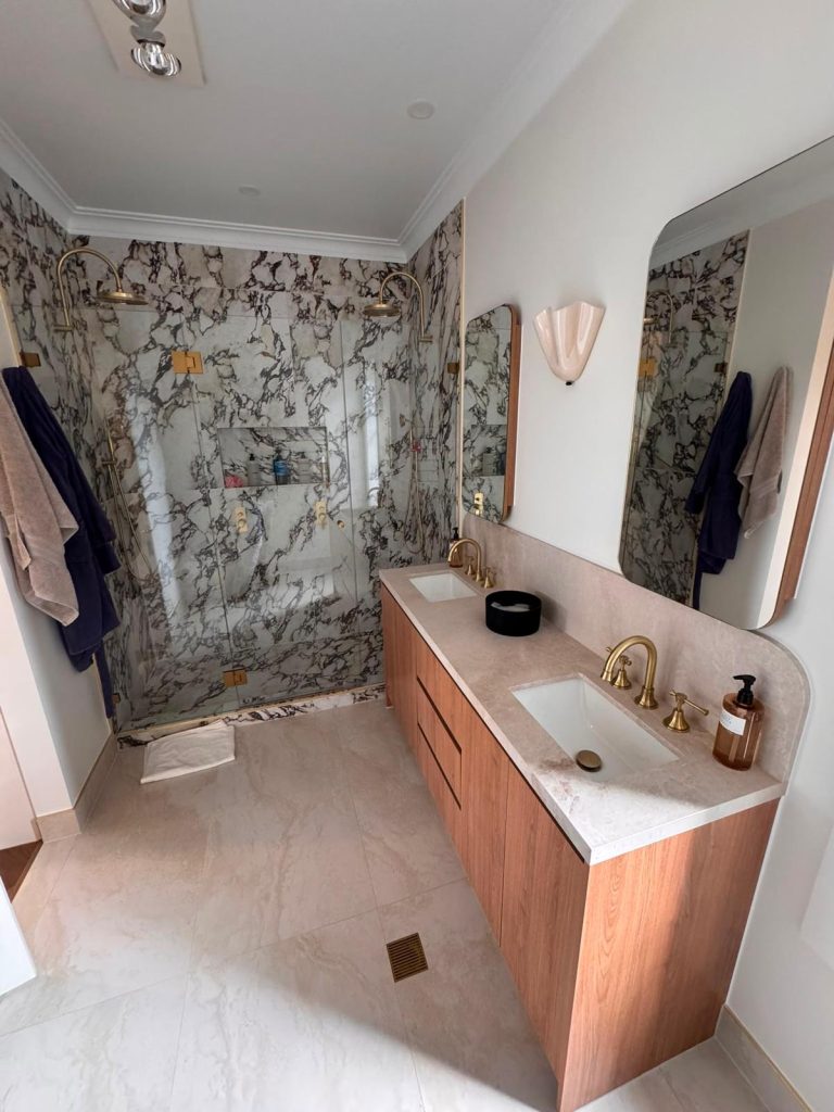 Bathroom with a timber wall-hung double vanity, stone top, brass tapware and a bold marble-tiled shower.