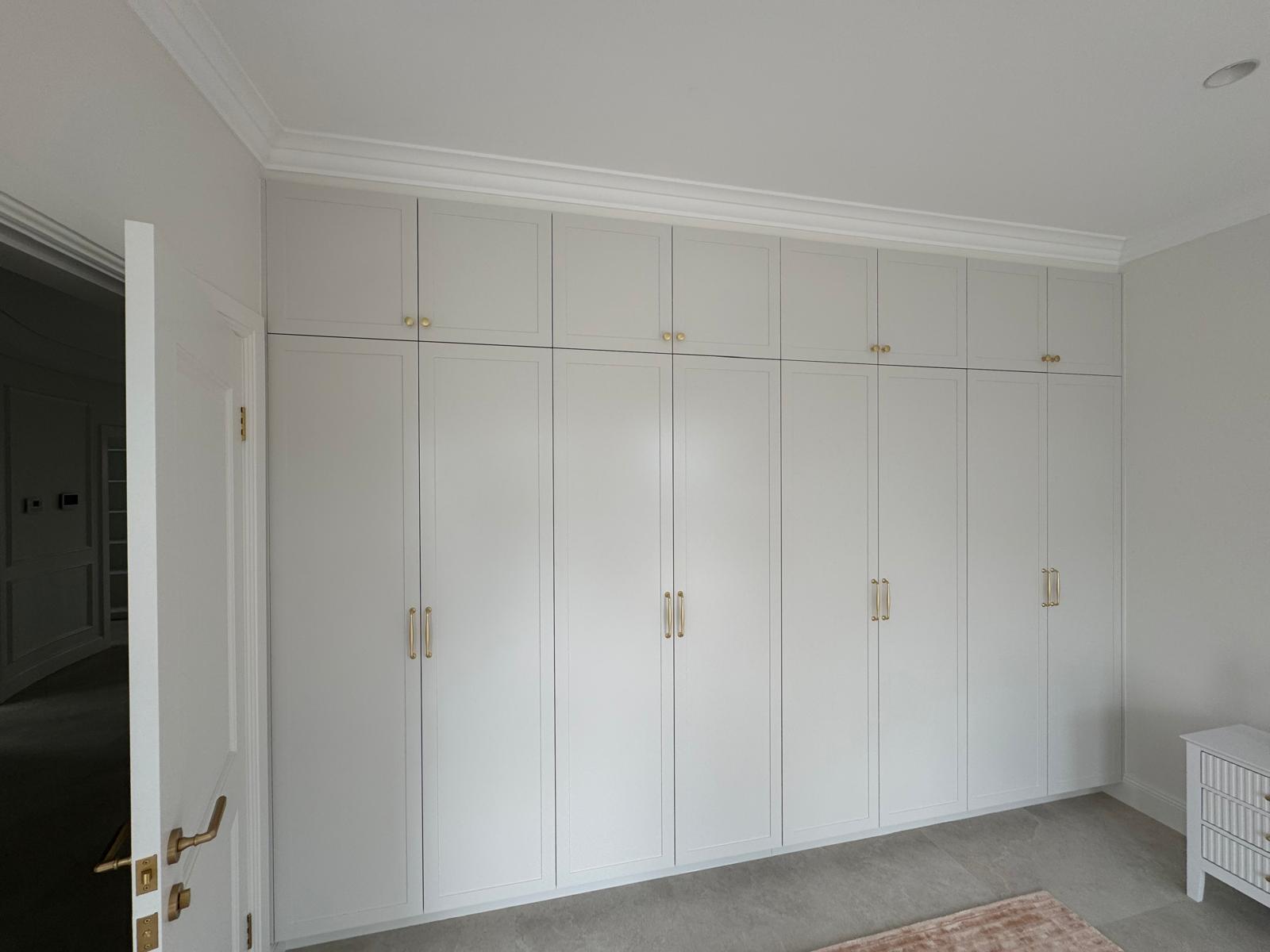 Bedroom wall of white shaker built-in wardrobes finished with crown moulding and brass pulls.