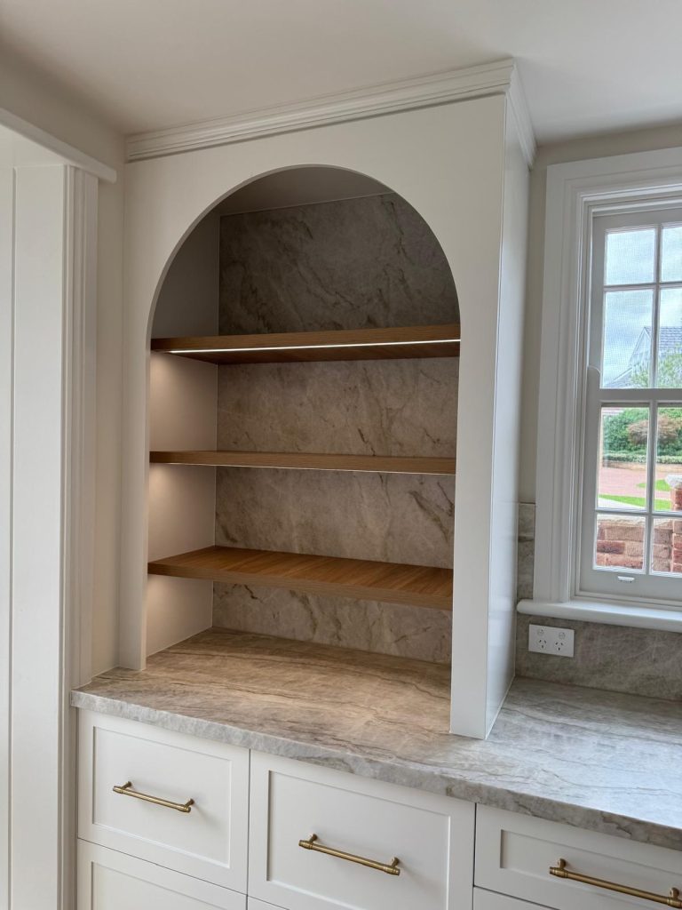 Arched kitchen niche with lit timber shelves and a stone backdrop above white shaker drawers.