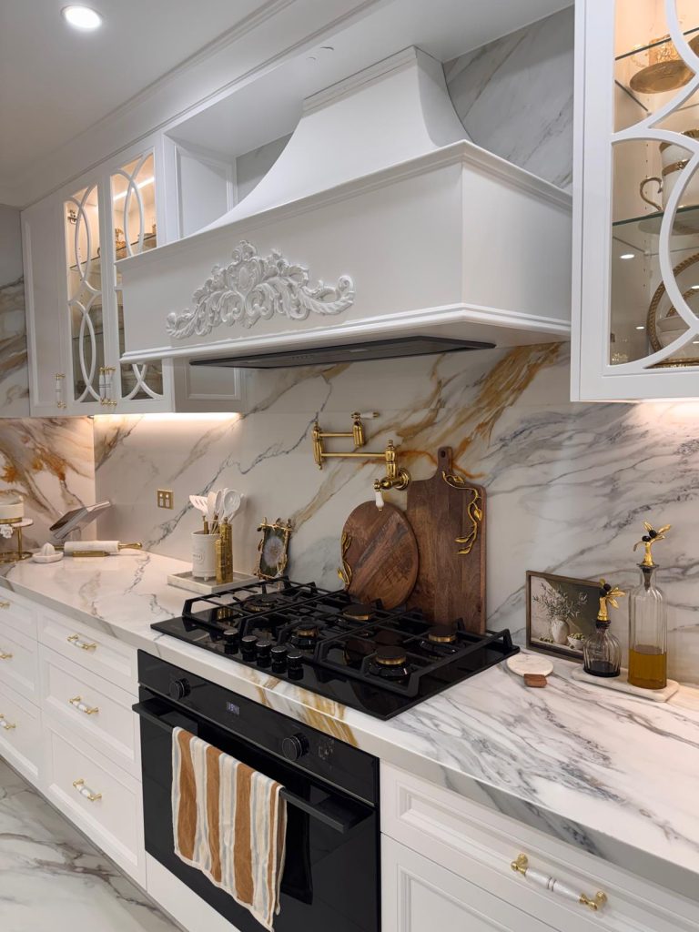Carved white canopy rangehood over a gas cooktop with a gold pot filler and marble-look splashback.