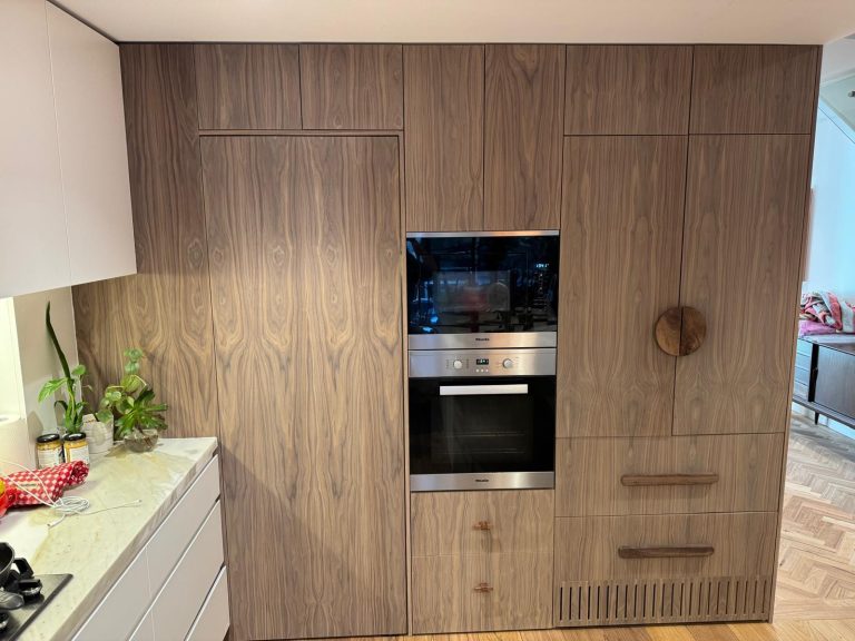 Wall of walnut-veneer tall cabinets with built-in ovens and timber pull handles.