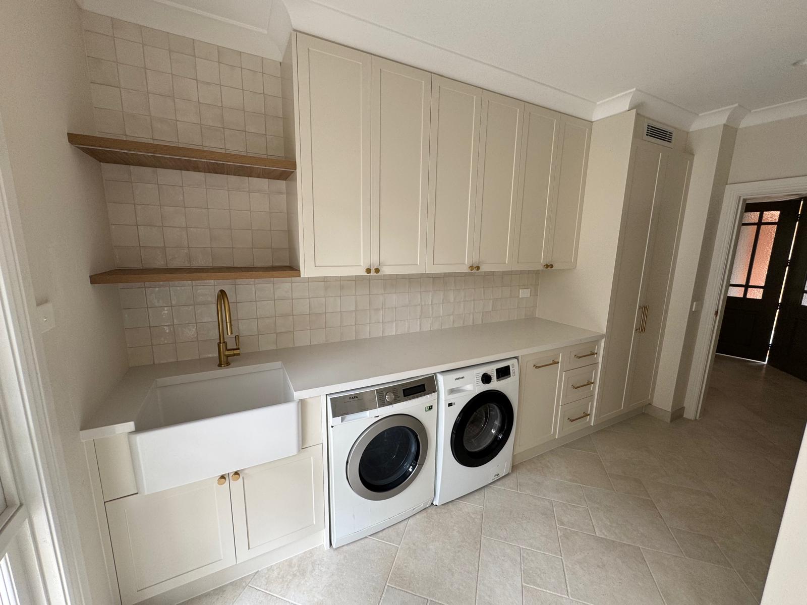 Laundry with a butler's sink, brass tap, timber open shelves and white shaker cabinets over the washer and dryer.